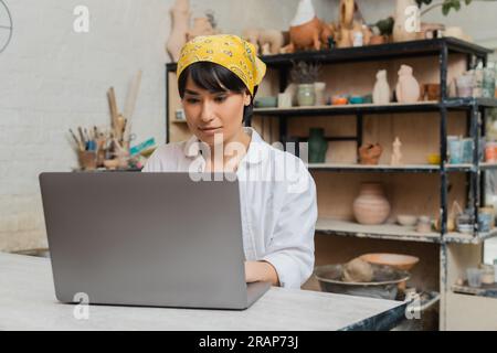 Young brunette asian female artist in headscarf using laptop at table while working in blurred ceramic workshop at background, pottery artist showcasi Stock Photo