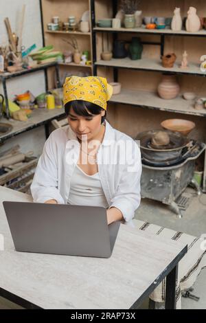 High angle view of young asian female artisan in headscarf and workwear using laptop while sitting at table and working in blurred ceramic workshop, p Stock Photo