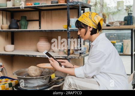 Side view of young brunette asian female artist in headscarf and workwear using digital tablet with blank screen while sitting in ceramic studio at ba Stock Photo
