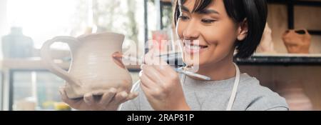 Smiling young asian female artisan painting on clay jug while working in blurred ceramic workshop at background, clay shaping technique and process, b Stock Photo