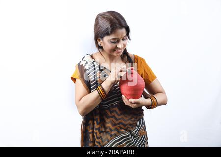 Happy rural Indian old lady in saree standing in white background inserting coin in the piggy bank. Aspirational lady showing saving money concept. Stock Photo