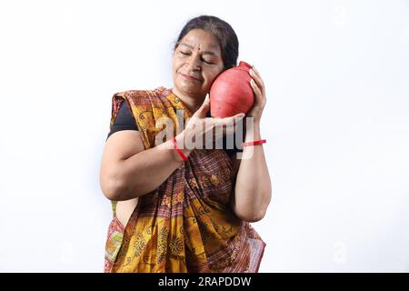Happy rural Indian old lady in saree standing in white background inserting coin in the piggy bank. Aspirational lady showing saving money concept. Stock Photo