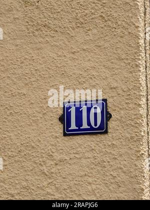 Number 110 house number plate. Blue plate with white numbers on a pale yellow building wall, no people. Stock Photo