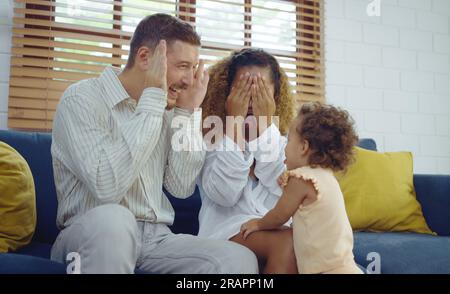 Happy cheerful parents and cute daughter are playing peekaboo on sofa together. Stock Photo