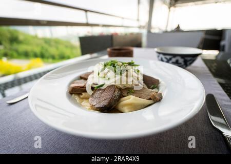 Composition of tasty Kazakh national dish beshbarmak in a plate with kazy, flour tortillas, onions on a table in a restaurant Stock Photo