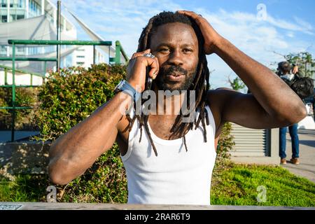 young black man with beard and dreadlocks of African ethnicity, sitting outdoors at sunset shocked and surprised, he is talking on the phone with one Stock Photo