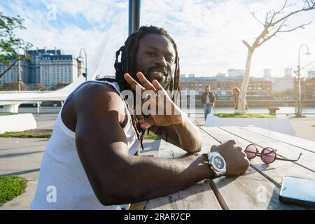 portrait of young tourist man with dreadlocks and beard sitting outdoors waving at the camera with his hand and smiling, sitting at a table at sunset Stock Photo