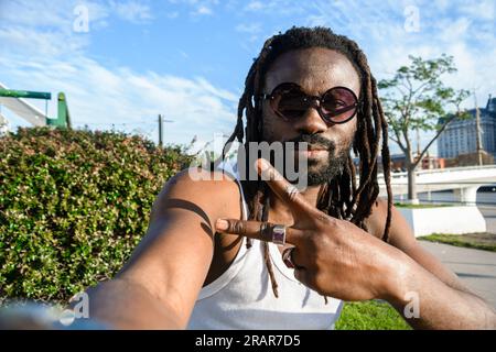 Selfie portrait of young African man with dreadlocks, glasses and beard sitting looking at the camera making v sign with hand and smiling with good at Stock Photo