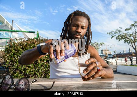 front view of young black man with dreadlocks, sitting outdoors at sunset, concentrated serving soda in a plastic cup to drink and refresh himself Stock Photo