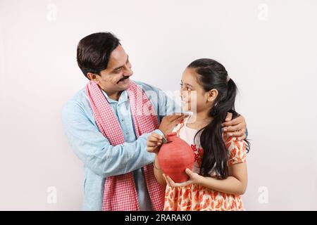 Happy Indian rural father and daughter holding piggy bank in hand, Saving money and banking concepts. Daughter inserting a coin into piggy bank. Stock Photo