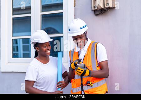 Two industrial contractors using mobile phone, standing and smiling outside an impressive building, representing teamwork and construction industry Stock Photo