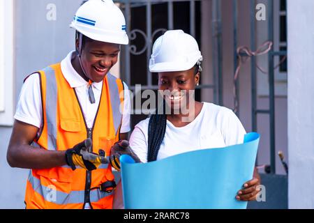 African male and female industrial engineers looking at blueprints while standing in front of a building project Stock Photo