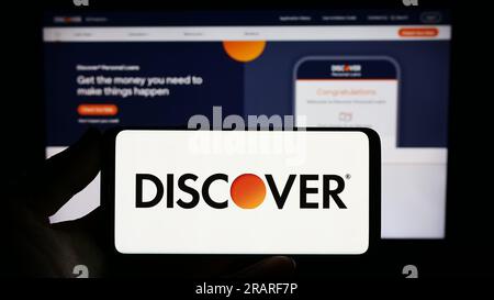 Person holding smartphone with logo of US company Discover Financial Services on screen in front of website. Focus on phone display. Stock Photo