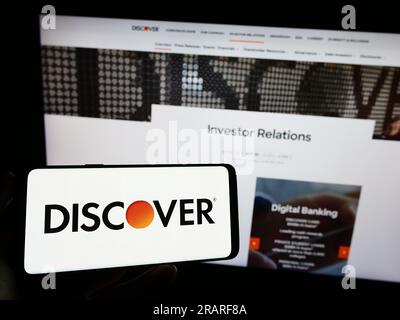 Person holding mobile phone with logo of American company Discover Financial Services on screen in front of web page. Focus on phone display. Stock Photo