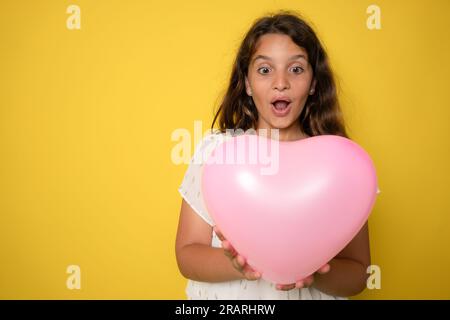 cheerful cute little lady receive birthday present hold heart shaped balloon isolated on yellow color background Stock Photo