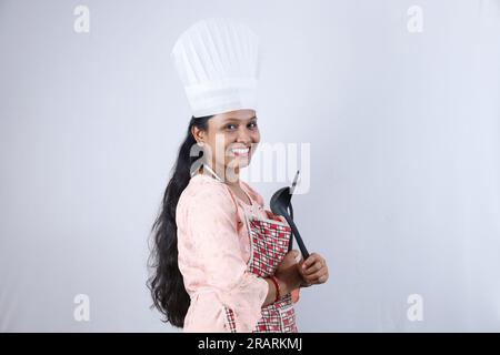 Happy Indian housewife holding frying spoon in hand and wearing a chef cap showcasing various moods of the house chef wearing apron. Stock Photo