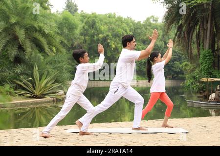 Trainer exercising and teaching kids the yoga poses in green environment early morning in park to maintain healthy lifestyle. International yoga day. Stock Photo