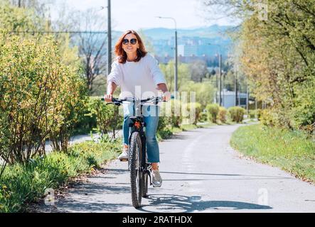 A happy smiling Caucasian woman in fancy sunglasses riding a bicycle on the city park street. Ecology sustainable transport and happy people concept i Stock Photo