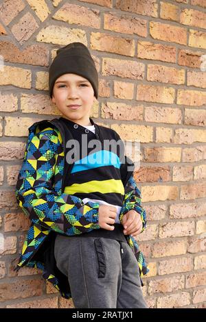A teenager in fashionable clothes stands leaning on a brick wall. Casual teenage fashion. Youth fashion and lifestyle. Stock Photo