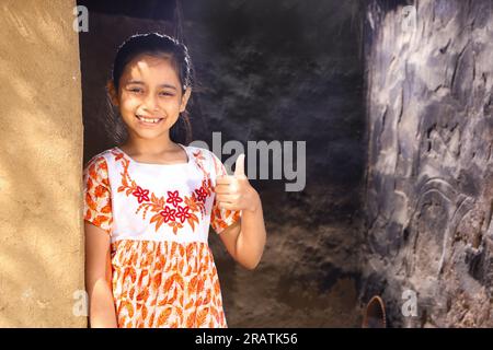 Happy rural Indian girl standing on her cottage gate wearing a gown, smiling and looking into camera in day time summer. Thumbs up with smile. Stock Photo