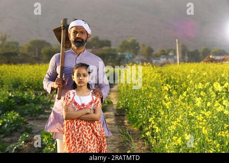 Beautiful dramatic portrait of Indian rural angry farmer and daughter standing in the mustard field wearing kurta pajama holding shovel in hand. Stock Photo