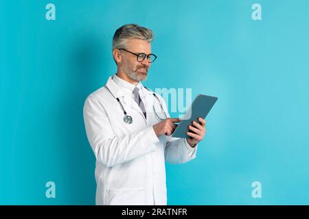 Happy middle aged doctor using digital tablet, blue background Stock Photo