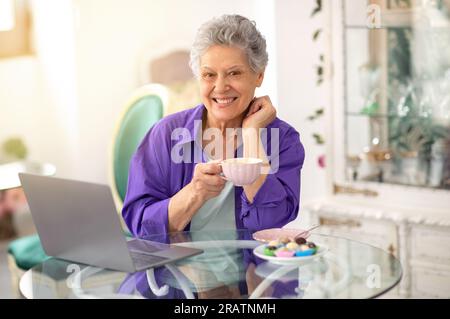 Happy Senior Lady Using Laptop And Having Coffee In Cafe Stock Photo