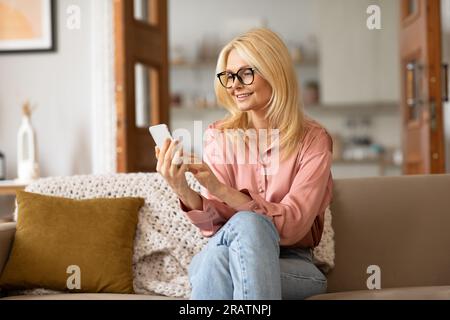 Happy Senior Woman Wearing Glasses Using Smartphone In Living Room Stock Photo