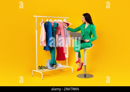 Happy woman fashion designer with laptop looking at clothes on warbrobe rack, using laptop computer, yellow background Stock Photo