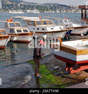Man paints a wooden boat on a summers evening by the Bosporus shoreline in Istanbul. Passenger ferries and the Asian side in the background. Turkey Stock Photo