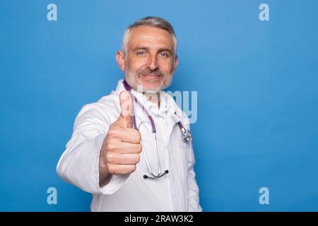 Good-looking cheerful handsome grey-haired middle aged man in medical workwear doctor showing thumb up and smiling at camera, isolated on blue backgro Stock Photo