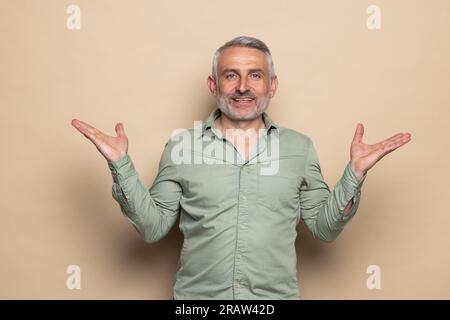 Elderly confused man shrugging shoulders looking puzzled spread hands isolated on beige background studio portrait. Stock Photo