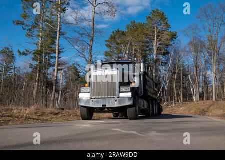 A black gravel truck and trailer parked on asphalt paved street, with trees and blue sky. Stock Photo