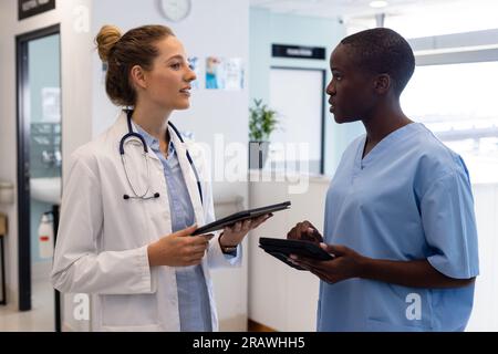 Diverse female doctors using tablets and talking in corridor at hospital. Hospital, communication, medicine and healthcare services, unaltered. Stock Photo