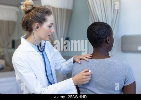 Diverse female doctor and patient examining patient, using stethoscope at hospital. Hospital, medicine and healthcare services, unaltered. Stock Photo
