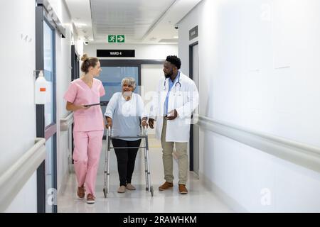 Diverse doctors and senior female patient with walking frame, walking in corridor at hospital. Hospital, medicine and healthcare services, unaltered. Stock Photo