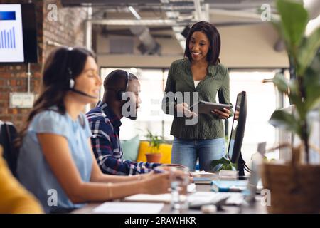 Diverse business people sitting at desks and using phone headsets and tablet at office. Casual business, communication and work, unaltered. Stock Photo