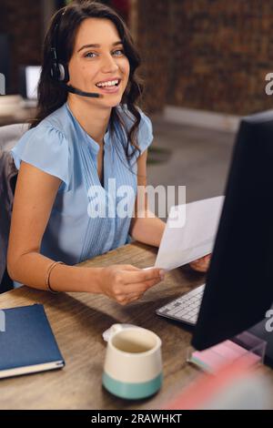 Portrait of happy caucasian businesswoman using phone headset and using computer, smiling at office. Business, confidence, communication and work, una Stock Photo