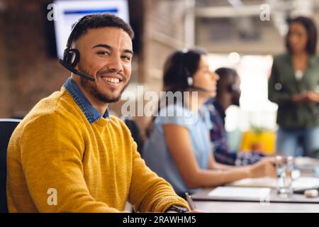 Portrait of happy biracial businessman using phone headset and smiling at office. Business, confidence, communication and work. Stock Photo