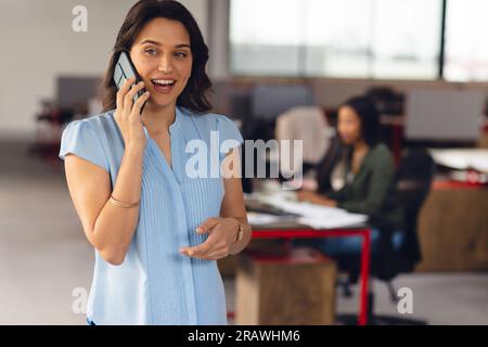 Happy caucasian businesswoman talking on smartphone, with colleague in background at office. Casual business, communication and work, unaltered. Stock Photo