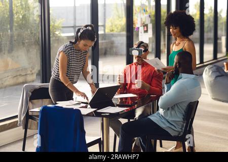 Diverse casual business colleagues in discussion, using vr headset and laptop at office meeting. Casual business, teamwork, meeting, communication and Stock Photo