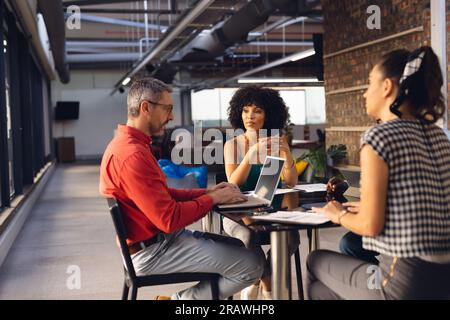 Diverse casual business colleagues talking and using laptop at office meeting. Casual business, teamwork, meeting, communication and work, unaltered. Stock Photo