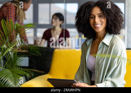 Portrait of happy biracial casual businesswoman on sofa, holding tablet and smiling at office. Business, communication and work, unaltered. Stock Photo