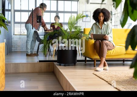 Diverse casual businesswoman using tablet and colleagues discussing work at office. Business, communication, teamwork and work, unaltered. Stock Photo