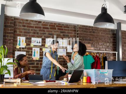 Happy diverse fashion designers using tablet and laptop, discussing project at desk in studio. Business, teamwork, communication, meeting, brainstormi Stock Photo