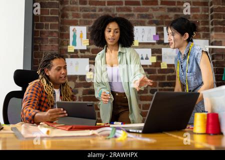 Diverse fashion designers using tablet and laptop, discussing project at desk in studio. Business, teamwork, communication, meeting, brainstorming, fa Stock Photo