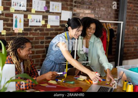 Happy diverse fashion designers using laptop, discussing project at desk in studio. Business, teamwork, communication, meeting, brainstorming, fashion Stock Photo
