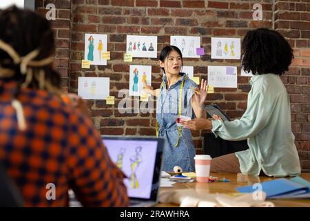 Happy diverse fashion designers using laptop and discussing designs in studio. Business, teamwork, communication, meeting, brainstorming, fashion and Stock Photo