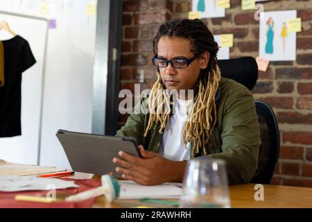 Focussed biracial male fashion designer sitting at desk using tablet at studio. Business, work, creativity, communication, fashion and design, unalter Stock Photo