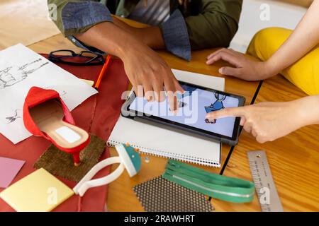 Midsection of diverse fashion designers using tablet discussing design sketches at studio. Business, teamwork, communication, fashion and design. Stock Photo